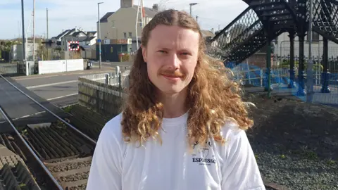 A man, with long, light brown curly hair and a light brown moustache. He is wearing a white t-shirt and is standing in front of a train track. 