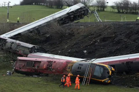 Owen Humphreys / PA Media A red and white train has derailed and is on its side, with each carriage sprawled at an different angle. The train tracks are atop a muddy mound in a grassy field. There are workers in high-vis uniforms dotted around the scene. Ladders have been placed on the train's front.