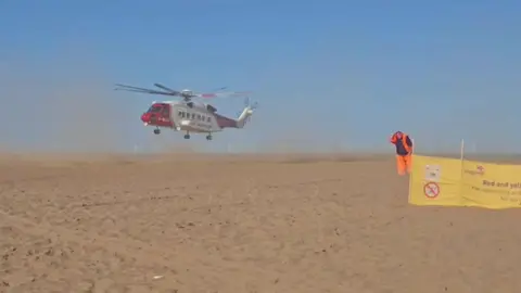 Supplied Sand is whipped up as a large red and white helicopter comes into land on a sandy beach under a blue sky. To the right, a man wearing orange hi-vis overalls bows his head as he stands next to a yellow Lifeguards sign.