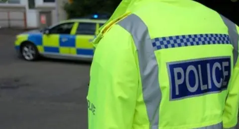The back of a police officer, in a high-vis police jacket. In the background is a police car with blue flashing lights. 