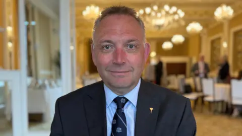 A white man in a dark blue suit jacket and dark blue tie with a pale blue shirt looking at the camera with a slight smile.