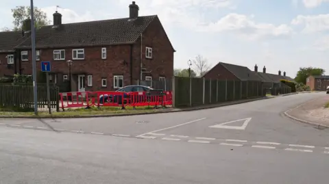 Martin Giles/BBC A road junction is shown with red brick houses to the left of a road, with red temporary barriers in place of some sections of wooden fencing.
