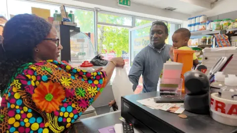 Nnenna Okonkwo wearing a bright and multi-coloured top and serving a customer who is wearing a shirt and jumper and holding his child.