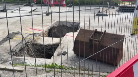 Two holes can be seen in concrete on the ground behind a metal fenced off area 