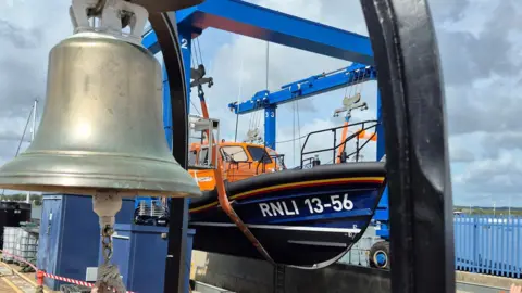 A large silver bell is in the top left-hand corner and behind it is an orange and blue RNLI lifeboat. It says RNLI 13-56 on it. It is suspended above the water on a crane. The sea can be seen in the distance.