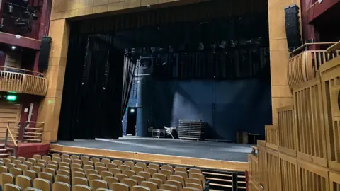 A theatre stage with seats in front made of a light coloured wood. Balconies on wither side of the stage in the same wood. 