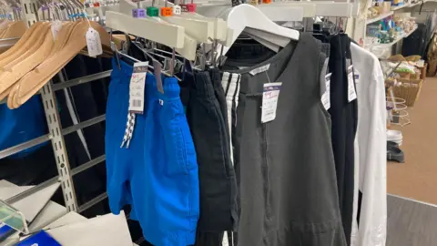 A clothing rack in the charity shop with grey school dresses, white shirts, and blue and black P.E shorts on coat hangers. They all have price tags of 50p