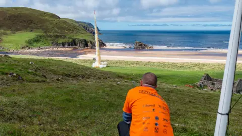 Ben Adams A member of Lura wearing an orange T-shirt crouches down as they watch the orange G2 rocket launch from an area of shoreline at Cape Wrath. The rocket leaves a trail of white smoke.