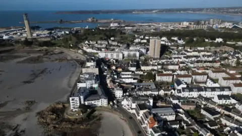 BBC A drone shot of Jersey. There are a multitude of different buildings, and the tide is out, leaving the muddy seabed beneath.