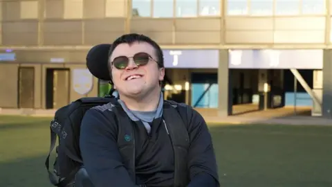 Tom Williams/BBC Man in a wheelchair wearing sunglasses and smiling on a football pitch.