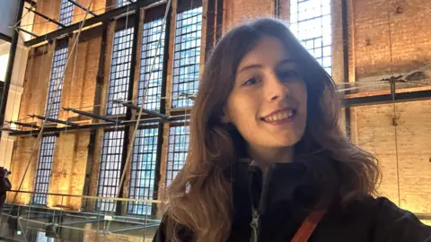 Emma Cook Emma is smiling for the camera, taking a selfie. She has long brown hair and behind her is a big bricked building with big windows and architectural features