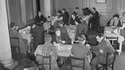 A black and white image of Queen's College, Taunton, 1943, Miss Pauline Craske (centre, facing camera) enjoys a meal at a British Restaurant in London. The restaurant is very well-patronised and many other people can be seen eating their lunch, circa 1943. (Photo by Ministry of Information Photo Division Photographer/ Imperial War Museums via Getty Images)