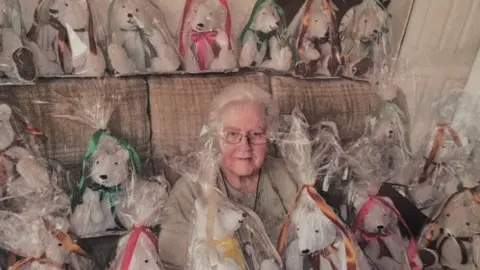 Family handout An elderly woman with short grey hair, glasses and a beige top. She is surrounded by teddy bears, grey in colour and all wrapped in polythene bags.