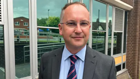 Leader of Herefordshire Council, Councillor Jonathan Lester. He is wearing a grey shirt, light blue shirt and dark blue tie with red strips and glasses. 