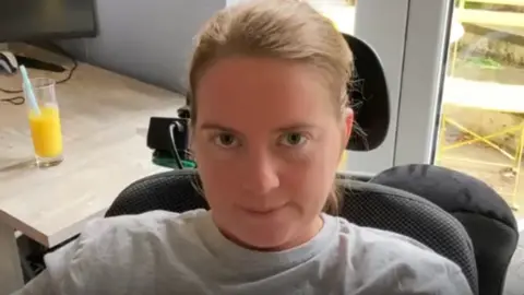 A woman with blonde hair looks directly at the camera. She is wearing a grey top and sitting in a chair with a head support. To the side of her is a table with a glass of juice on it.
