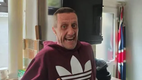 GMP A man in a mauve hoodie smiles while standing in a kitchen. He has short cropped brown hair.