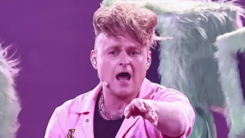 Sam Battle aka Look Mum No Computer performing on stage during Eurovision dress rehearsals in a pink jumpsuit pointing towards the camera