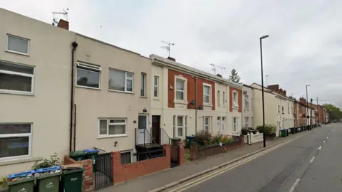 A Google image of Lower Ford Street in Coventry. The road has terraced houses on both sides in cream and red brick. There are steps leading up to some properties and many of the houses have bins in front of them.
