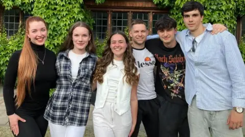A mixed group of young men and women stand smiling in front of a brick building, covered in ivy. They are dressed casually.