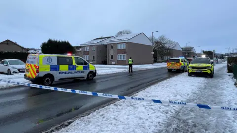 Several police cars can be seen parked along a road in a housing estate. Police tape has been put up to restrict access to the area. Snow is lying on pavements and rooftops and a police officer can be seen standing in the road. 