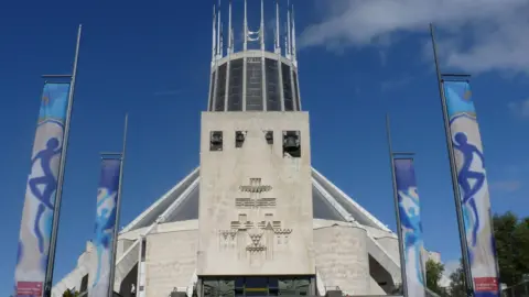 White stone tower of Liverpool Metropolitan Cathedral with engravings on stone front and banners showcasing an event at the sides.