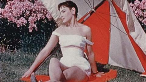 Brunette model with short hair, in a white short dress with left strap falling down, sitting on red towel on grass, with pink flowers in background, with a red and white umbrella positioned behind her.