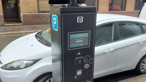 A black parking machine with a contactless panel on it. There's a white car parked on the side of the road behind it.