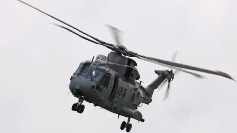 UK MOD Crown copyright The image shows a large grey military helicopter flying in the sky with its rotors spinning. The landing gear is down, suggesting it may be preparing to land or has just taken off.