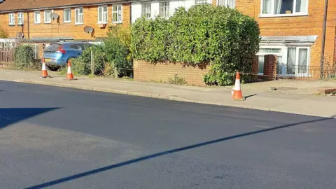 A section of Norcot Road resurfaced in the previous phase of improvements in March on a sunny day. A car is parked in front of one of the houses.