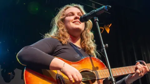 A young person, with long blonde hair, holding and playing a guitar, performing into a microphone. It looks like she is on a stage.