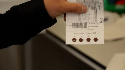 Someone holds a card showing results of a heel prick test which has four round spots of red on the bottom edge of the card.