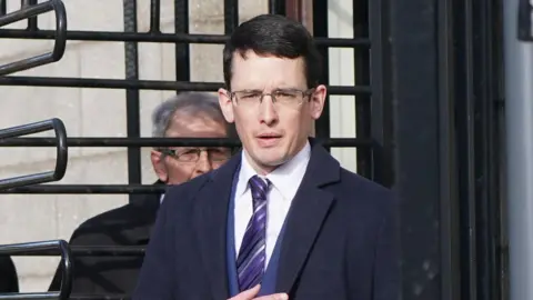 PA Media Enoch Burke, wearing a suit and tie, walks through a turnstile