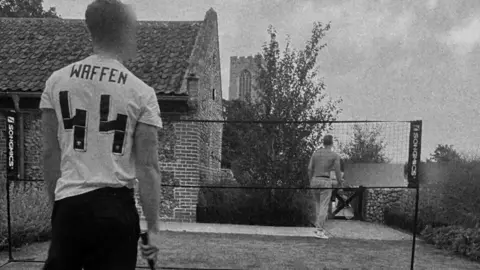 Telegram A member of Active Club, wearing a white shirt celebrating the SS, plays badminton with another shirtless member on grass outside a brick hall, close to a church