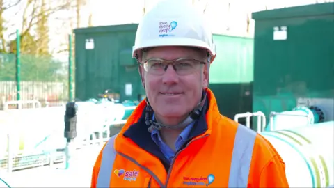 John Fairhall/BBC A man wearing a orange hi-vis jacket and a white helmet. He is also wearing a safety glasses and smiling at the camera.