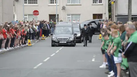 Pacemaker Three hearses drive down a road. On either side, large crowds of people watch. 