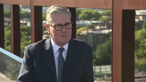 Sir Keir Starmer sat by a balcony overlooking part of London, with a bridge in the background. He is wearing a blue suit and blue tie.