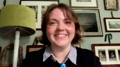 Morgan Ellis Leah wearing a black jumper and striped shirt smiles directly at the camera. Behind her is a wall of photo frames and a green lamp.