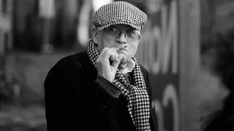 Christopher Furlong/Getty A man wearing a checked cap and scarf, plus glasses, draws on a cigarette and looks into the distance in this black and white shot.