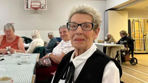 A woman with short hair is wearing black framed glasses and is wearing a white shirt and a black blazer. She is sitting at a table with a green gingham tablecloth and other people sitting around. 