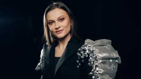 A dark haired girl, smiling at the camera, she is wearing a black blazer with silver ruffle detail and jewels on one shoulder