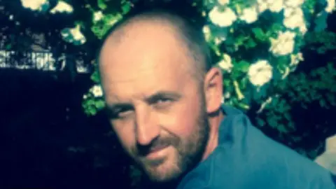 John Shelton smiling at the camera: a man with a brown beard and brown hair, pictured standing in a garden, wearing a blue shirt.
