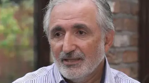 Consultant surgeon Dr Nasser Kurdy with grey hair and beard wearing a white and black striped shirt. He is indoors, sitting in front of a window with bare brick wall behind him.