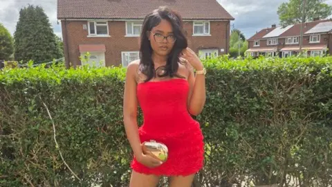 Handout Erica De Sousa Correia wears a red dress and holds a gold handbag. She wears glasses and has voluminous dark hair. She is posing for a photo outside in front of a tall hedge. There are houses in the background.