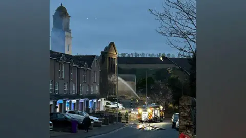 PA Media Fire crews battling a fire at a disused church, with a jet of water going into the building, area starting to get dark, fire engine lights can be seen.