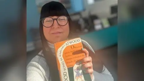 East Sussex County Council A woman with a black fringe and glasses holds the Good Bones book, which is orange with black writing on.