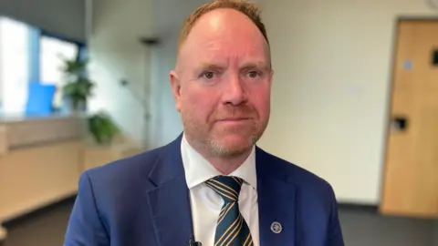 Andrew Judd, CEO of National Association of Funeral Directors has short red hair and a beard, he wears a striped tie, white shirt and smart blue jacket and is pictured in an office. 