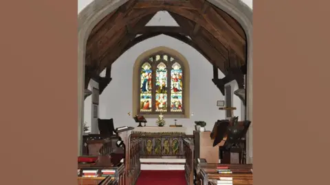 Lynda Poulter/Geograph Interior of St Andrew's Church, Greensted, looking towards the altar. It shows an arched stained glass window above a cloth-covered altar. It has white walls and the a pitched timber roof. There is a red carpet on the nave and wooden pews can be seen on either side.