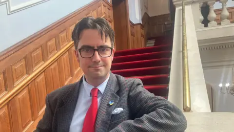 Simon Finlay A man with short brown hair and glasses smiling at the camera. He wears a jacket with a white shirt and a red tie.