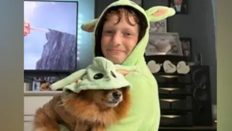 South Yorkshire Police A boy in a green Shrek hoodie holding a dog which is also in a green shrek outfit - they are in a living room of a house with a TV in the background and the boy is smiling.