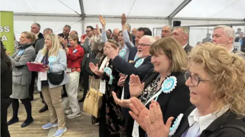 A group of people wearing blue Reform UK rosettes cheer and applaud in a large marquee. 